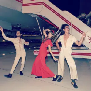 Three people in retro-style outfits strike dramatic poses on an airport tarmac at night, standing in front of a plane with red and white stripes.